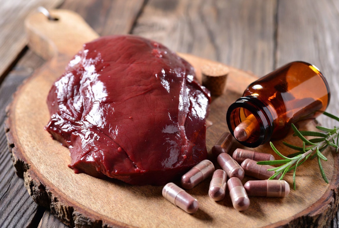 Desiccated beef liver supplement capsules spilled from an amber bottle beside a piece of raw beef liver on a wooden board