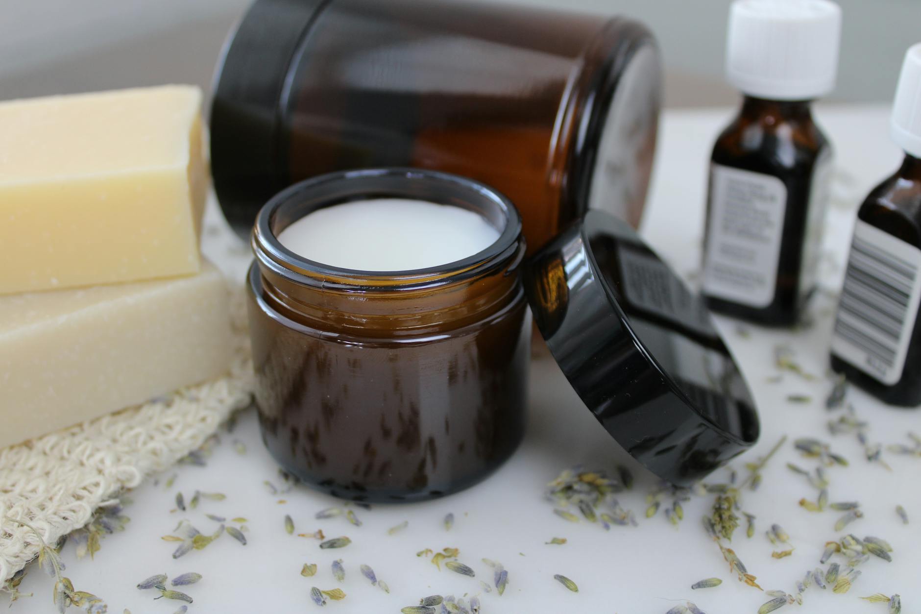 An open amber glass jar of creamy white rendered balm on a clean counter, surrounded by handmade soap bars, small essential-oil bottles, and dried lavender