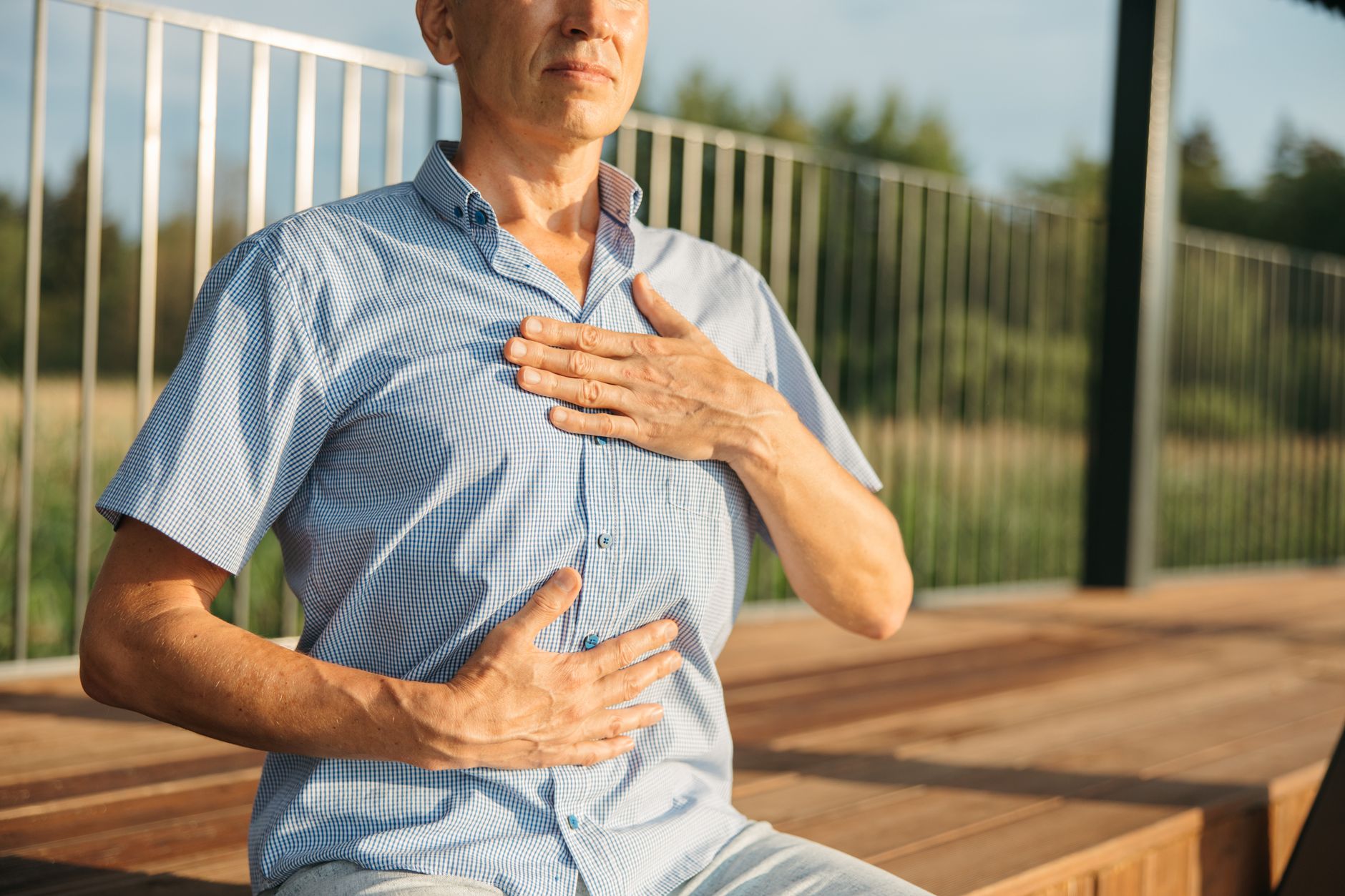A man sitting outdoors in morning light with eyes closed, one hand on his chest and one on his belly, practicing a slow diaphragmatic breathing exercise