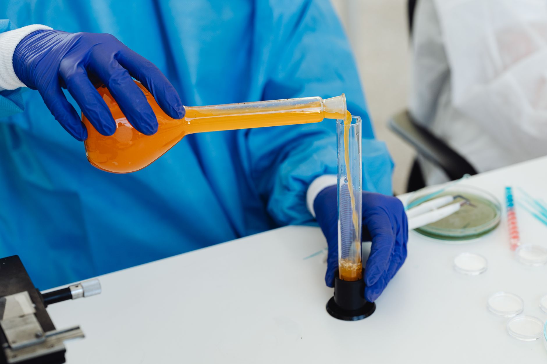 A gloved researcher pours an orange solution from a round-bottom flask into a test tube at a laboratory bench, with a microscope and petri dishes nearby, representing early-stage preclinical research