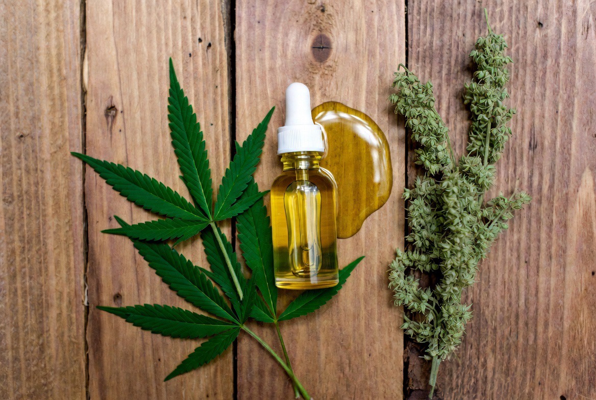 A glass CBD oil tincture bottle with its dropper resting beside fresh green hemp leaves on a neutral surface, showing the plant source of the extract