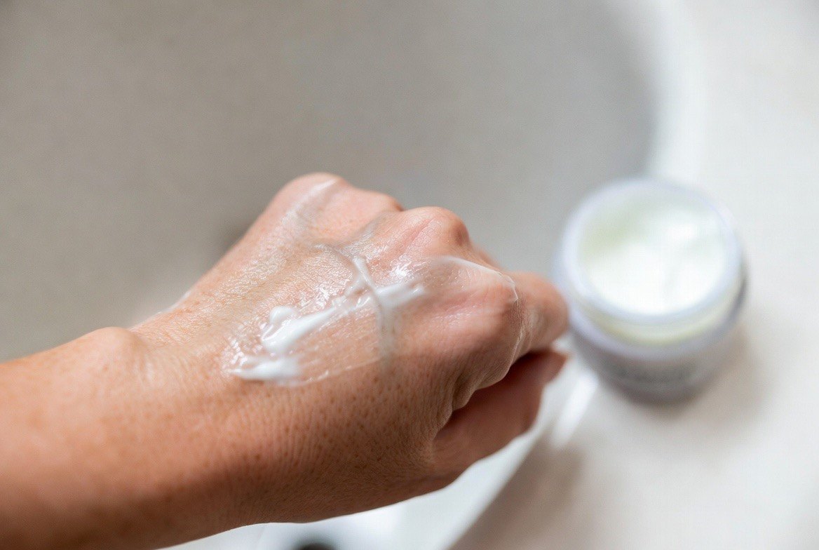 A person smoothing a rich, opaque white ceramide moisturizing cream across the back of one hand, the thick cream texture visibly spread and catching soft light on the skin