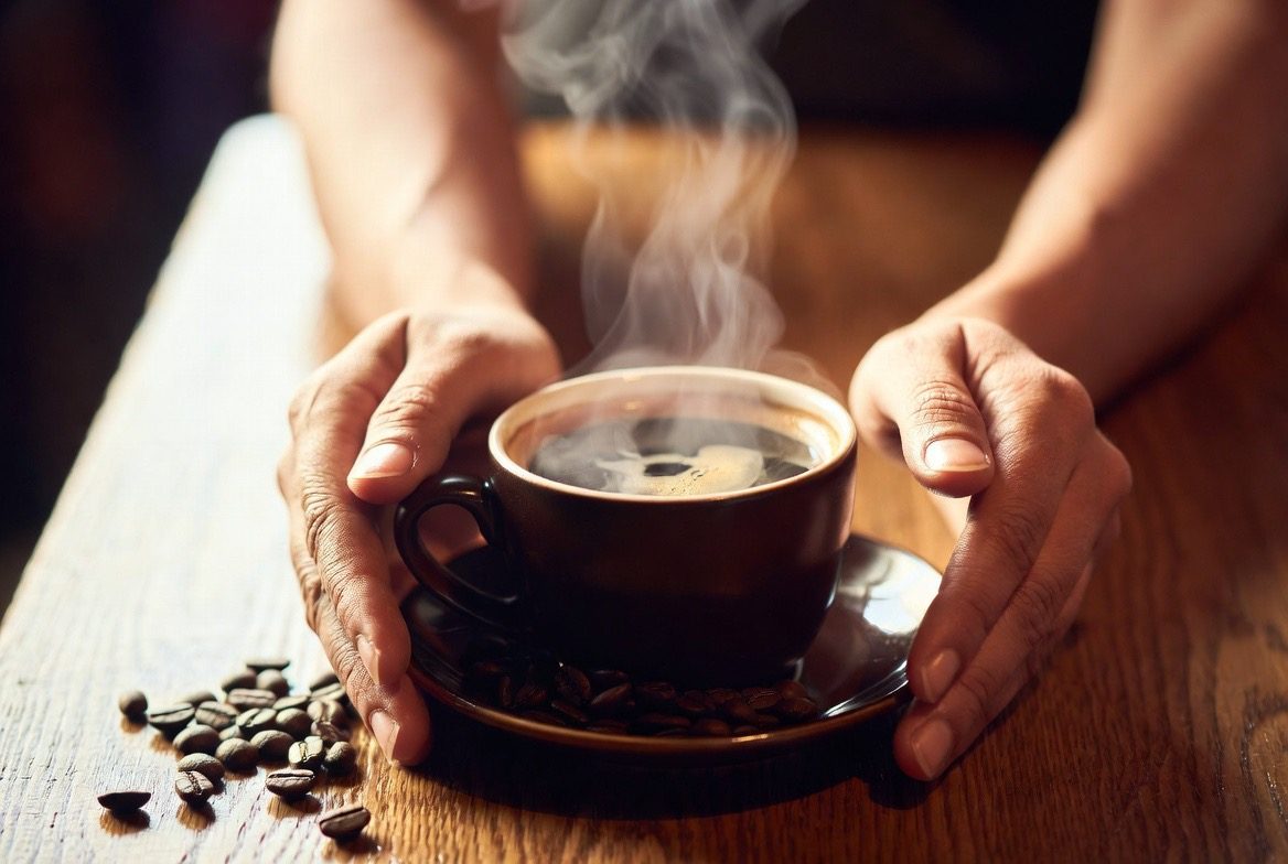 A person's hands cradling a steaming cup of black coffee on a saucer with scattered roasted coffee beans on a wooden cafe table in warm morning light