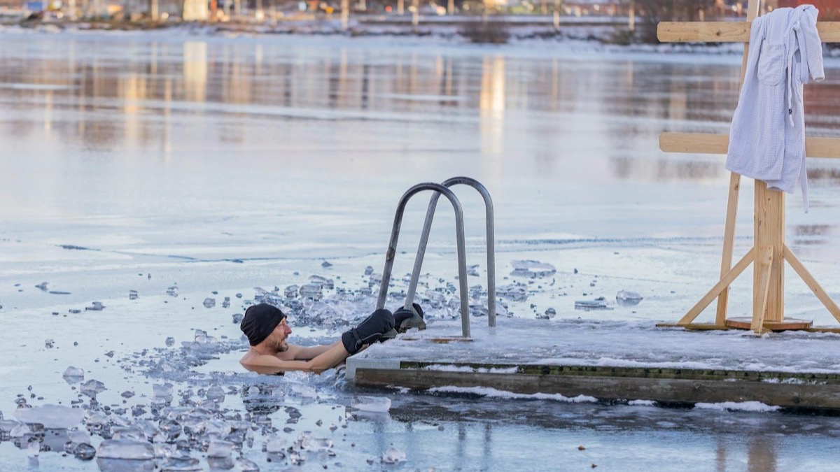 A man in a wool cap immersed in broken-ice cold water beside a dock ladder, robe hanging nearby — an outdoor cold plunge in near-freezing conditions