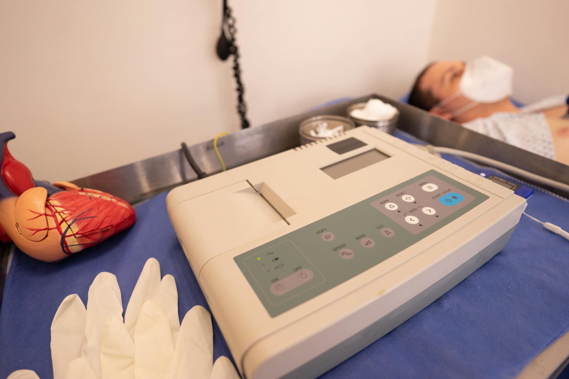 ECG machine and anatomical heart model on an exam table beside a resting patient — cardiovascular monitoring in a clinical setting