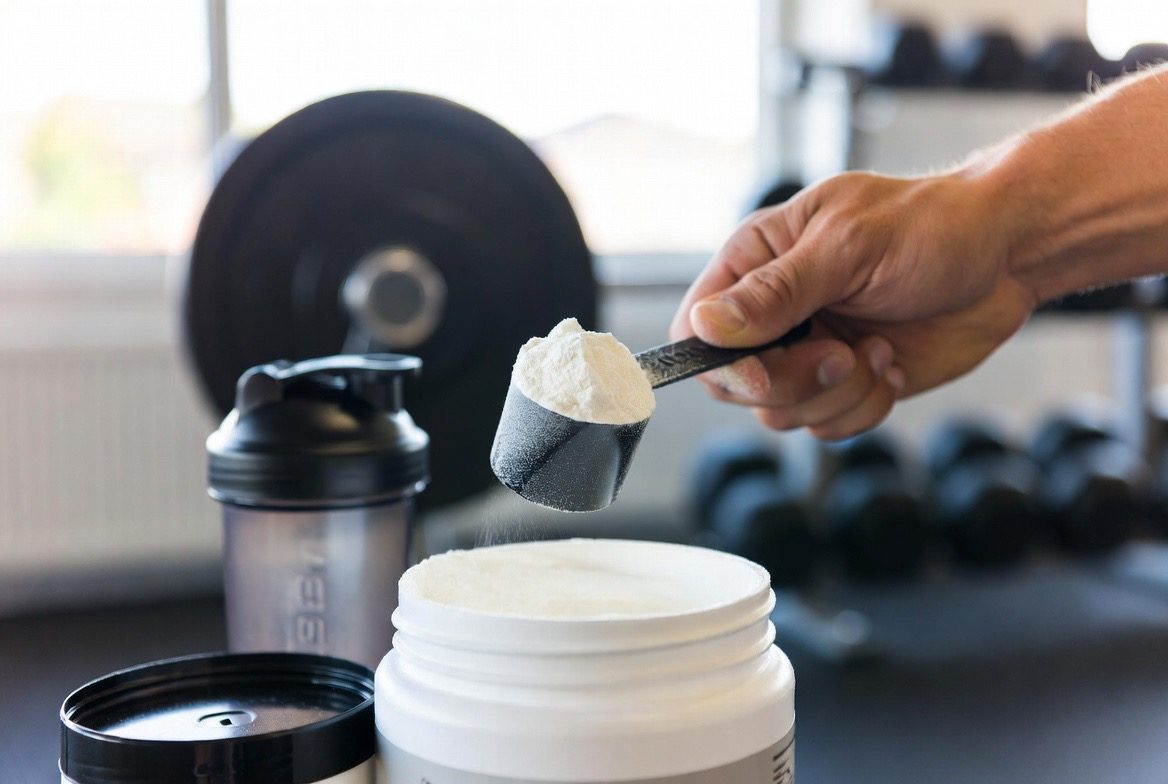 A hand lifts a heaped black scoop of white creatine monohydrate powder from an open tub, with a protein shaker bottle, a loaded barbell and dumbbells blurred in a gym behind