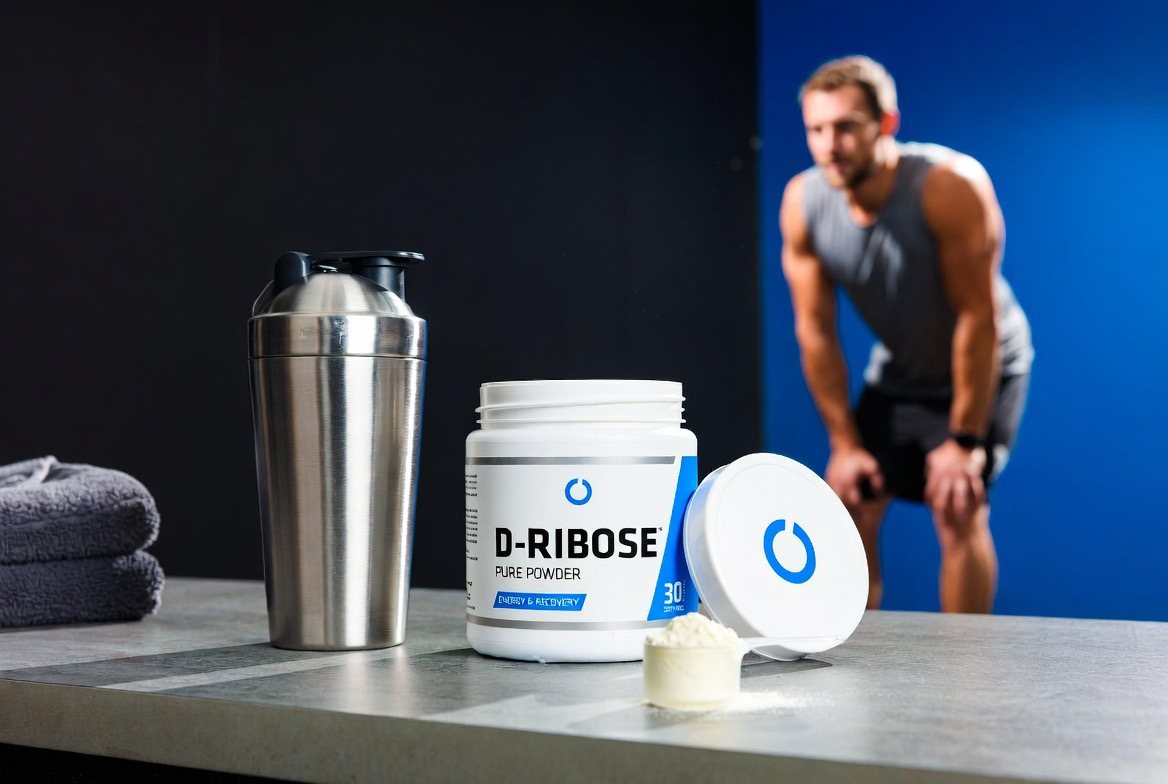 An open tub of white D-ribose powder with a measuring scoop resting on a kitchen counter beside a glass of water, the fine crystalline supplement sold as an ATP energy booster