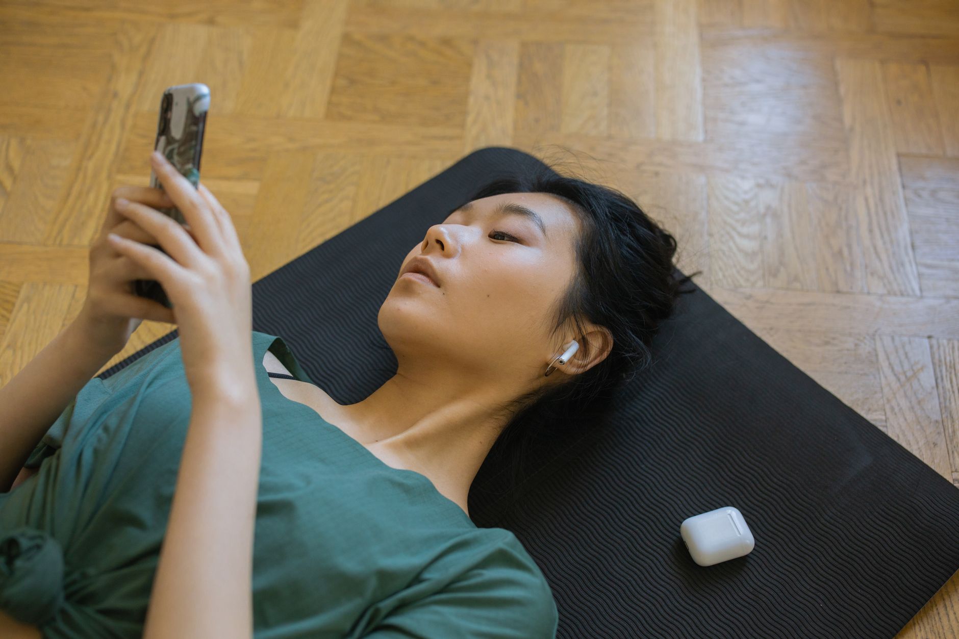 A young person lying back on a yoga mat with earbuds in, absorbed in a smartphone held above their face