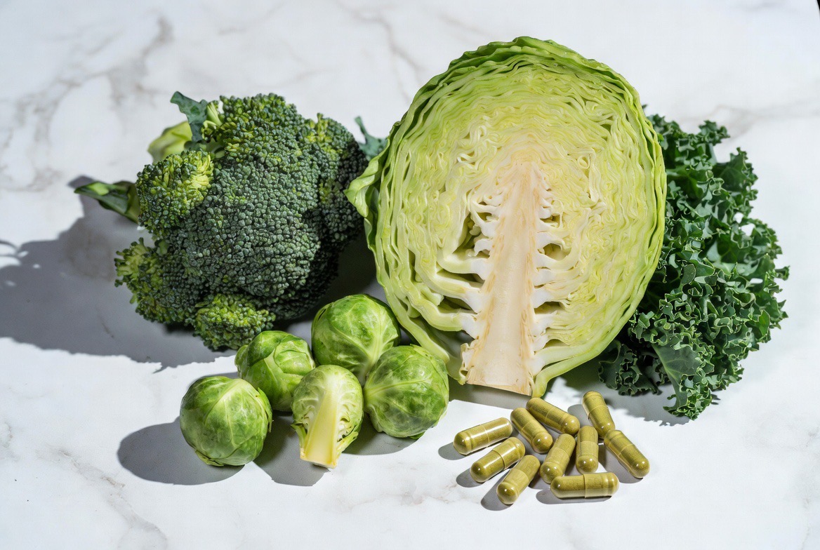 Fresh broccoli, cabbage, Brussels sprouts and kale — the cruciferous source of DIM — beside green supplement capsules on a marble surface