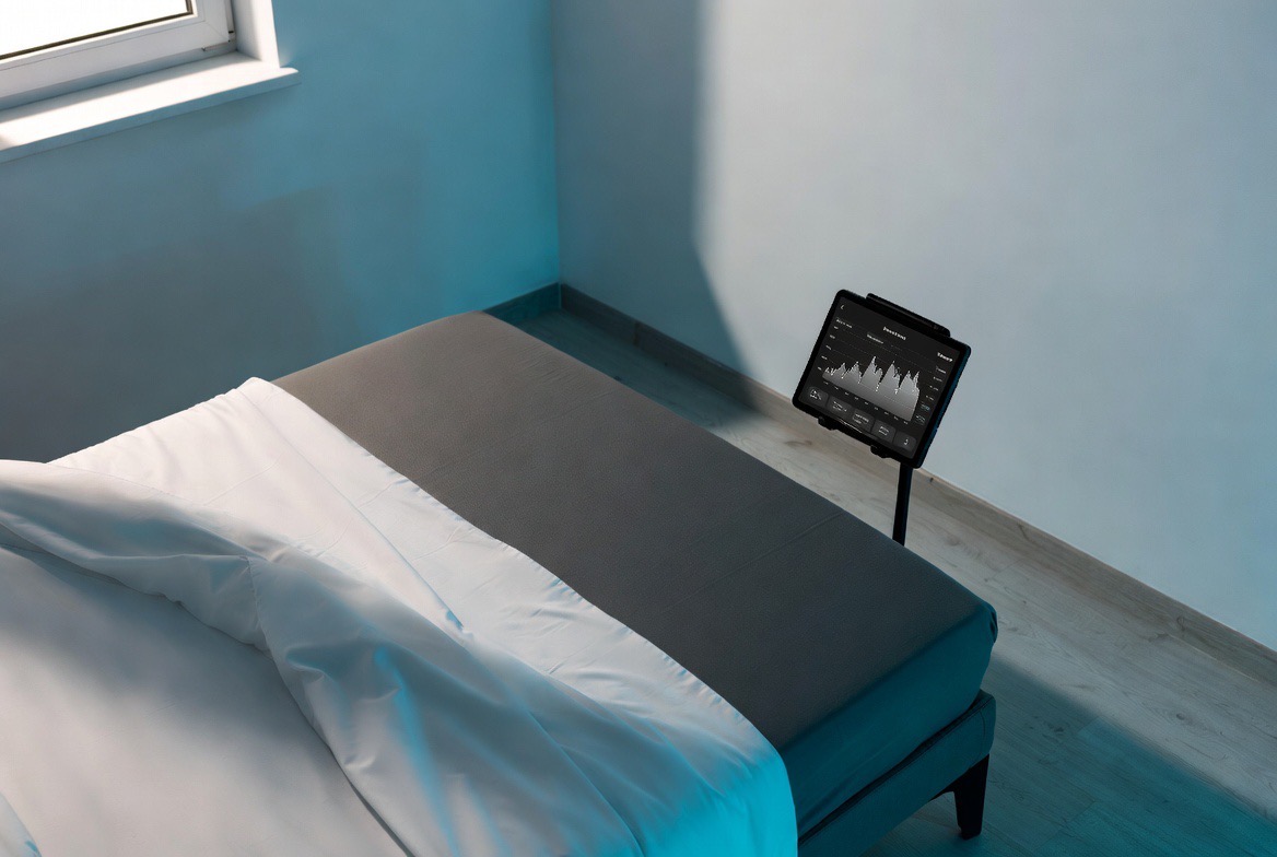 A neatly made bed fitted with a thin temperature-controlled mattress cover, its water tubing visible at the edge, beside a cool-toned bedside display showing a night of sleep data in a blue-lit bedroom