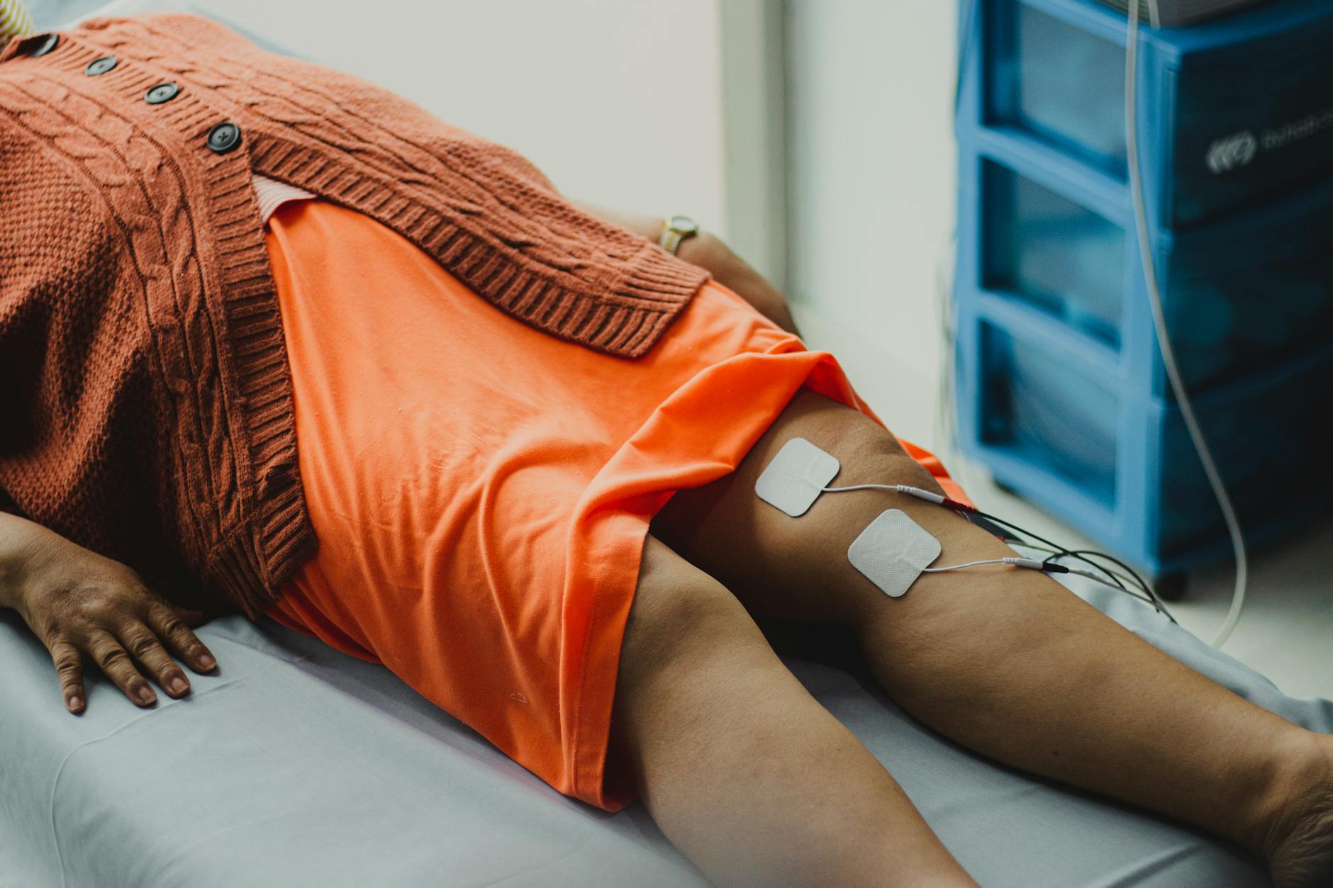 Two square electrode pads with lead wires placed on a reclining person's thigh, connected to an electrical stimulation unit in a clinical setting