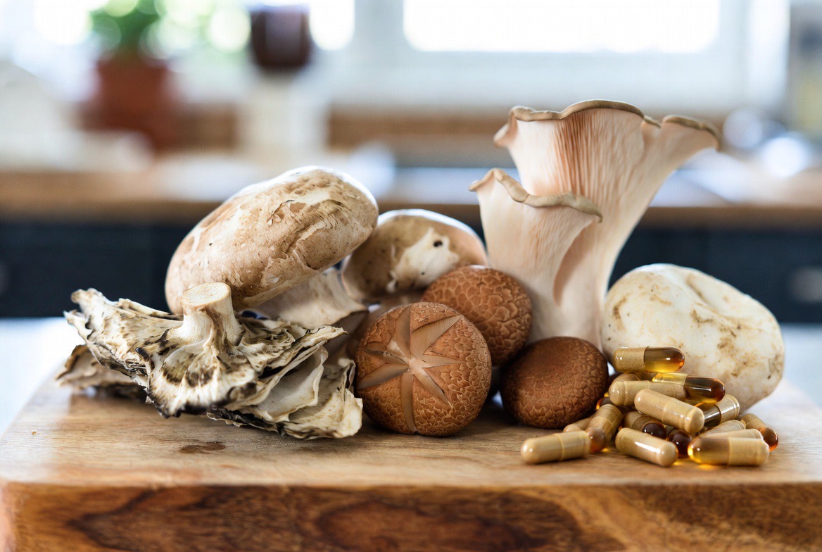 A spread of fresh whole mushrooms — the richest dietary source of ergothioneine — arranged on a neutral editorial surface beside a small dish of supplement capsules