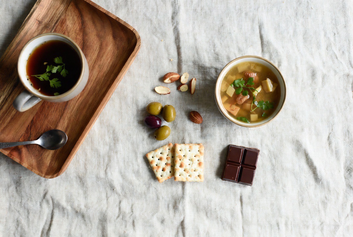 A small portioned plant-based fast-with-food spread &mdash; a tiny bowl of vegetable soup, a few olives and nuts, and a cup of herbal tea on a pale table, signaling the deliberately low-calorie fasting-mimicking diet; no people