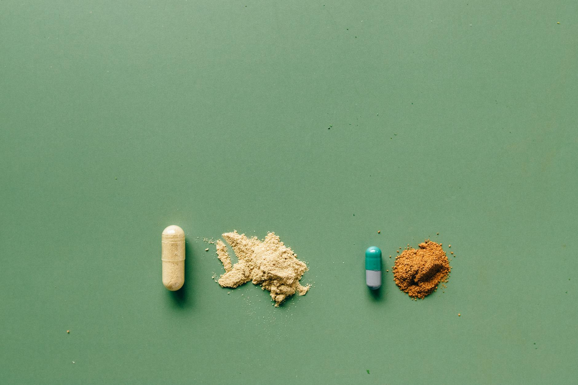 White and teal supplement capsules beside small piles of beige and orange supplement powder spilled on a calm green surface