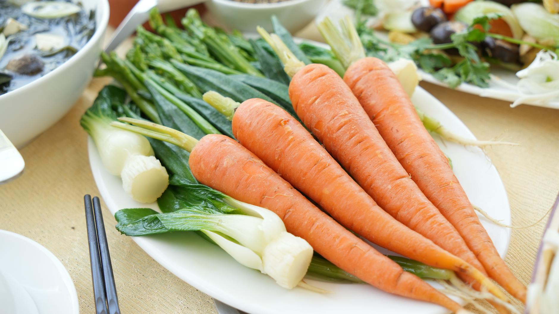 A white plate of fresh carrots and leafy green vegetables on a wooden table in soft natural light