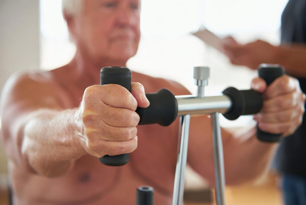An older adult squeezing a handheld hydraulic hand-grip dynamometer during a strength test while a clinician records the kilogram reading &mdash; the simple, cheap measurement that predicts mortality in large cohort studies