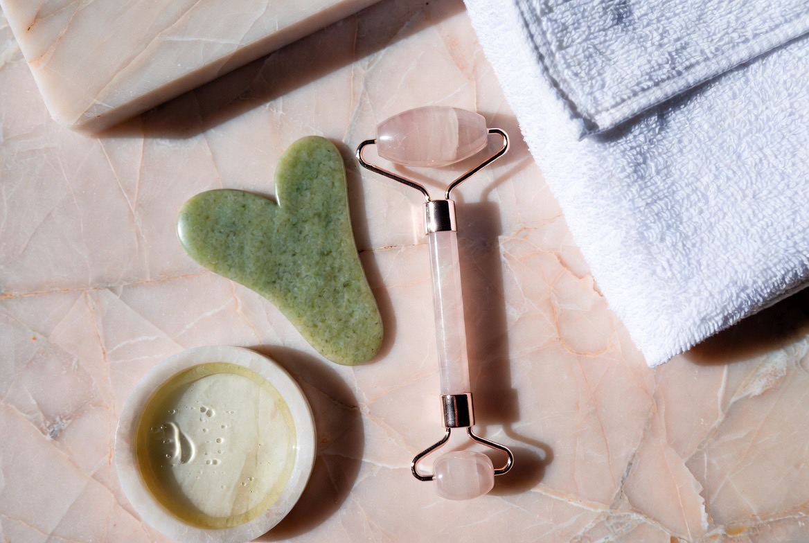 A jade gua sha stone tool and a rose-quartz facial roller resting on a clean minimalist vanity beside a small dish of facial oil