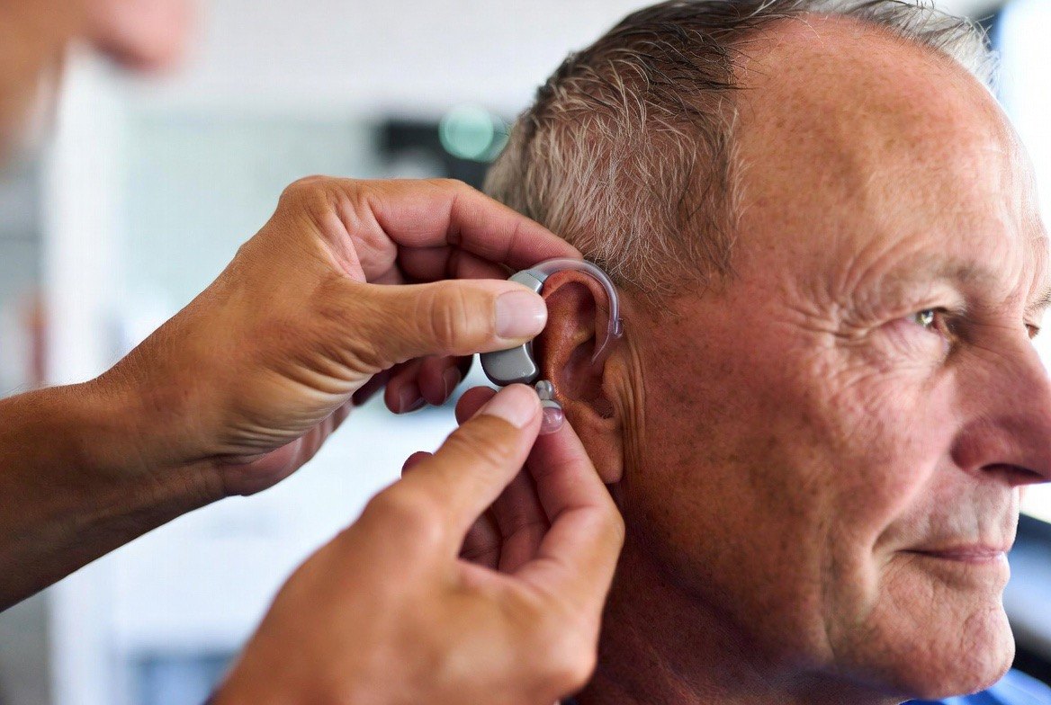 An audiologist fitting a small modern behind-the-ear hearing aid onto an older adult's ear during a clinic appointment &mdash; the low-risk intervention at the center of the hearing-loss-and-dementia question