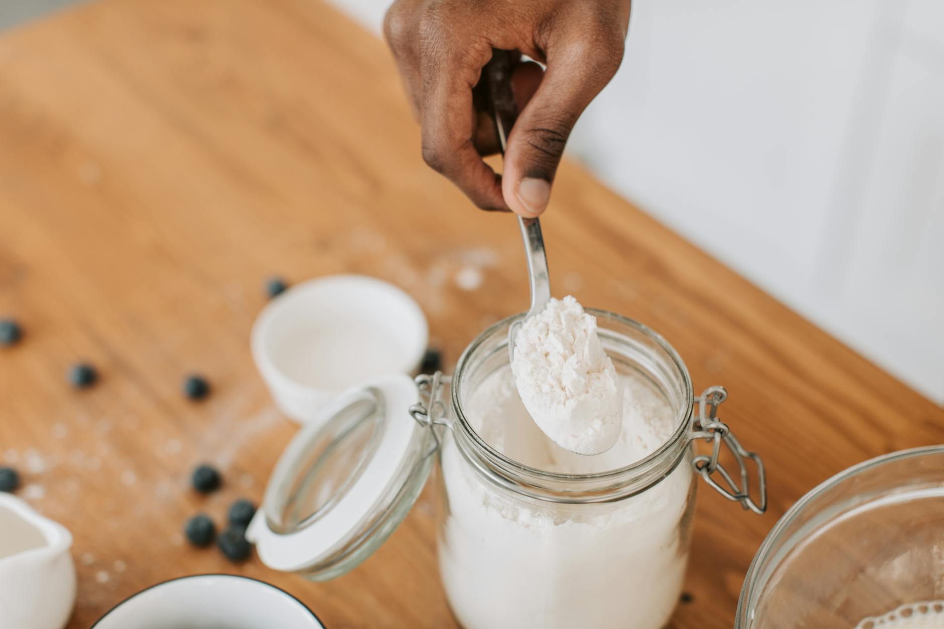 A hand scooping a spoonful of fine white powder supplement from an open glass jar on a wooden kitchen counter