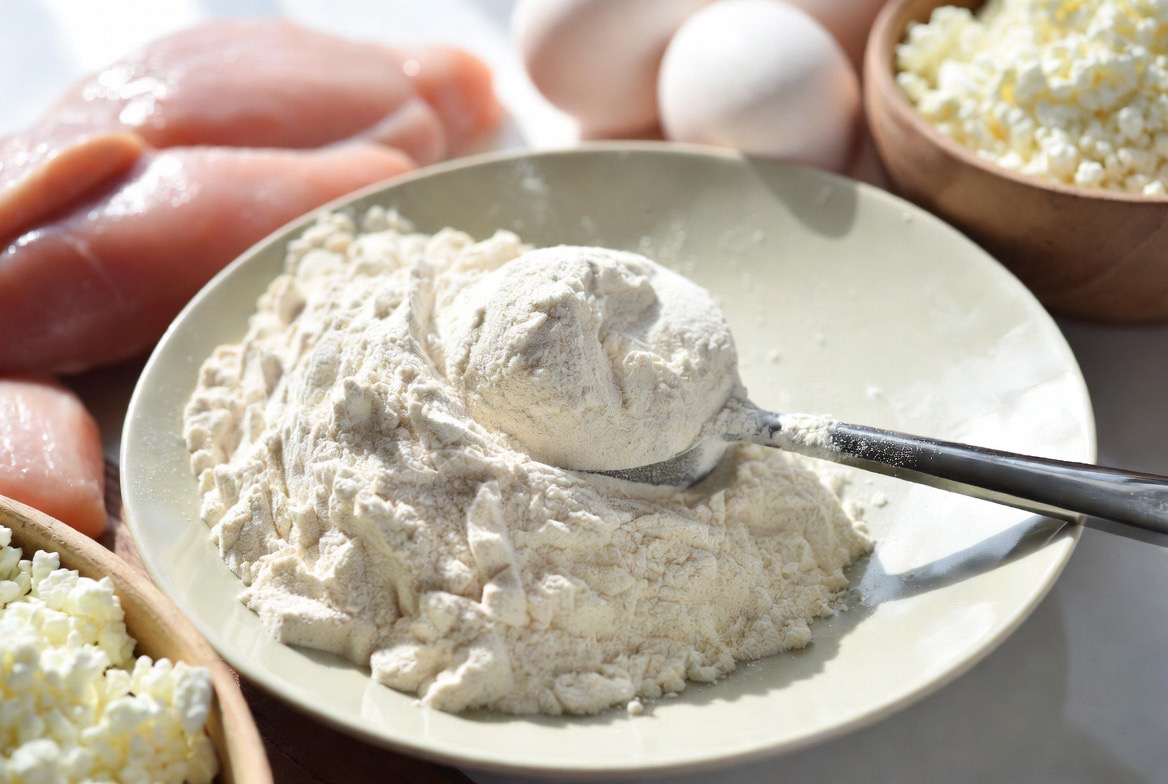 A scoop of white L-glutamine amino-acid powder on a plate surrounded by protein-rich glutamine food sources — raw chicken breast, eggs, and cottage cheese — in bright editorial light
