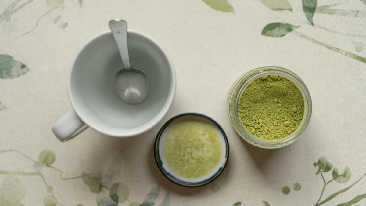A cup of green tea with loose green tea leaves scattered on a clean neutral surface in soft natural light &mdash; green tea is the dietary source of L-theanine