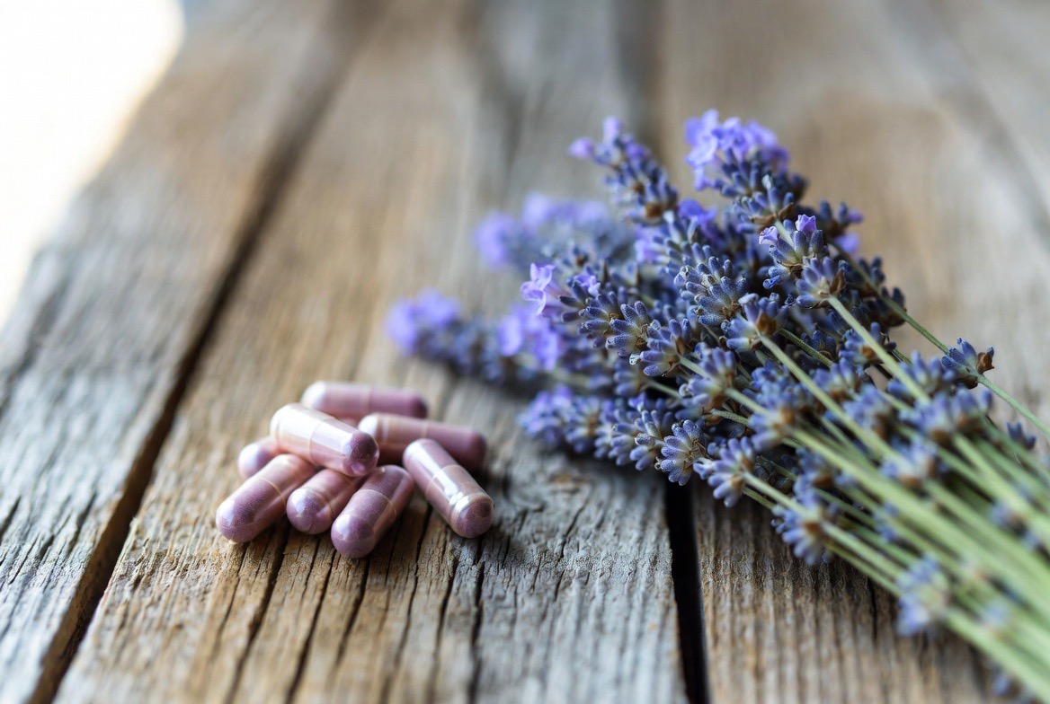 Fresh purple lavender flowers and sprigs arranged prominently beside a small cluster of lavender-colored capsules on a weathered wooden surface in soft natural light
