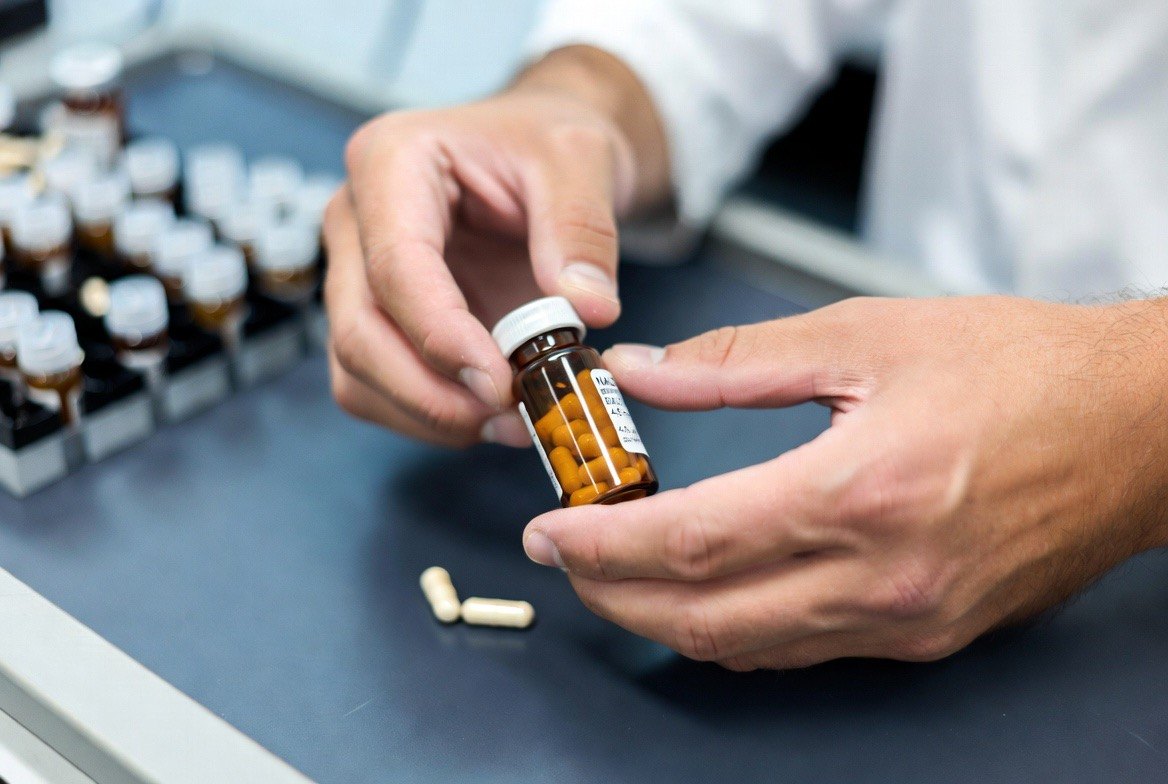 A pharmacist's hands at a compounding bench holding a prescription vial of small low-dose naltrexone capsules beside a labelled prescription, illustrating that LDN is a compounded, prescription-only preparation rather than an off-the-shelf supplement
