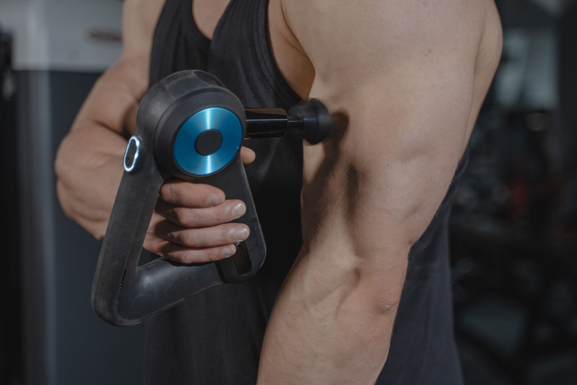 A person in a gym holding a handheld percussion massage gun against their upper arm and shoulder muscle