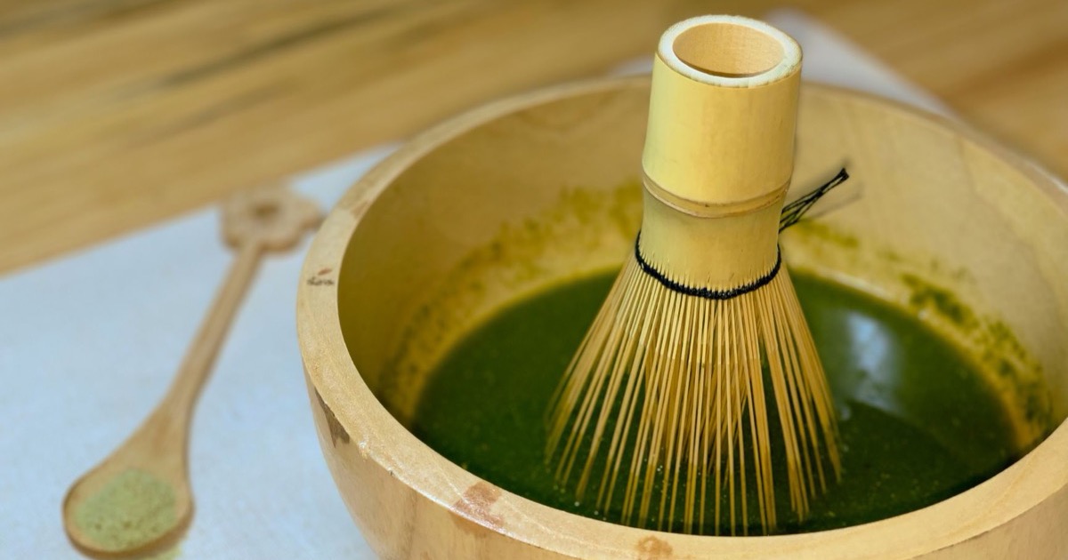 Bright green matcha whisked in a wooden bowl with a bamboo whisk, beside a wooden spoon of green matcha powder on a light surface in soft natural light