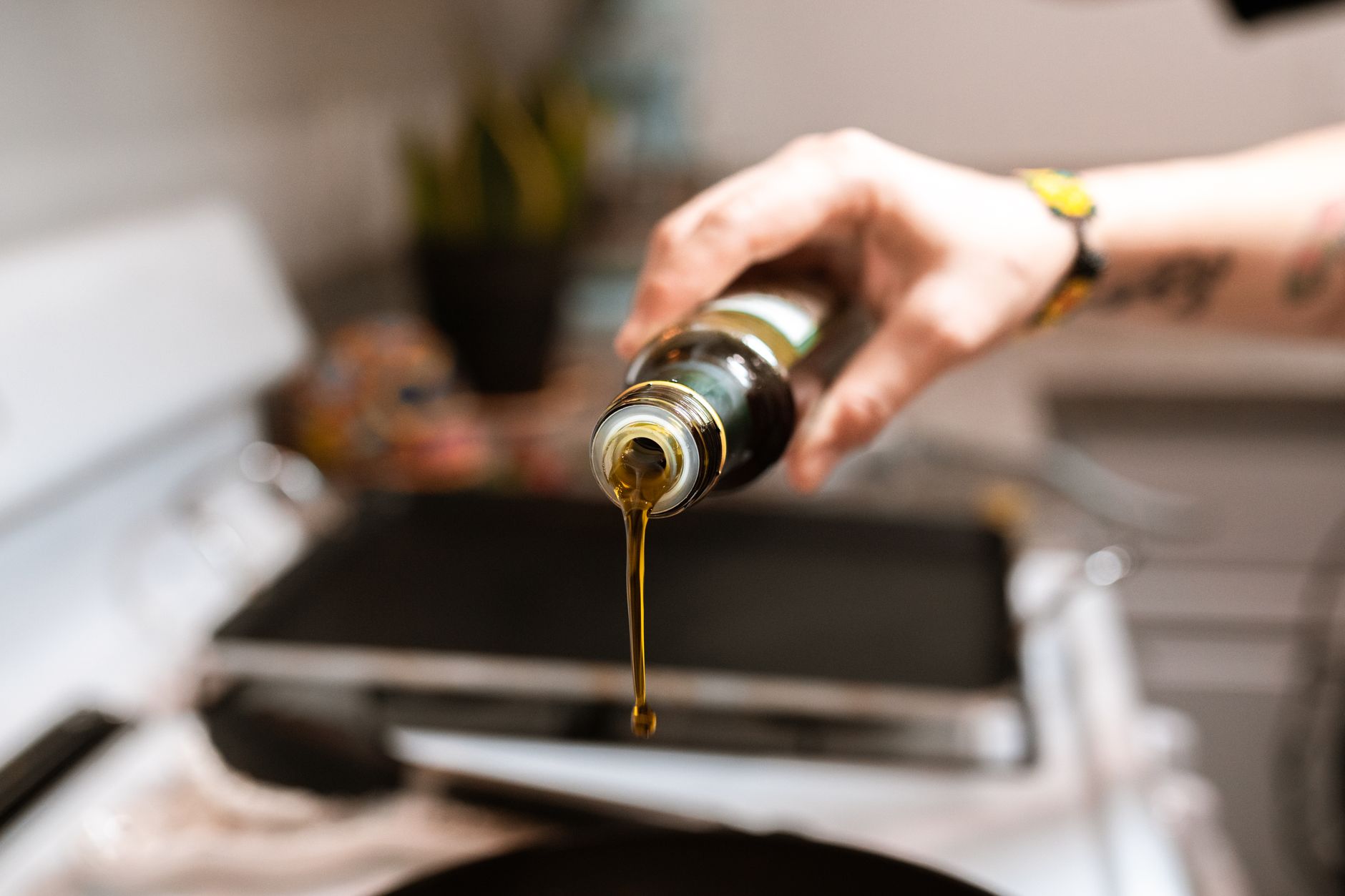 A hand pouring golden dietary oil from a bottle into a pan on a kitchen stovetop, representing the cooking-and-coffee oils MCT is marketed alongside