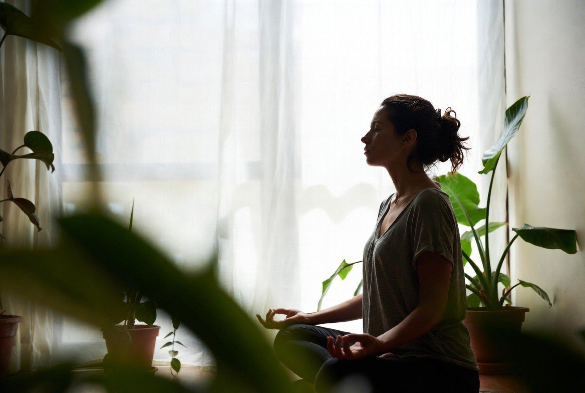 A woman sitting cross-legged in an upright meditation posture, eyes closed and shoulders relaxed with hands resting on her knees, silhouetted against a bright curtained window with houseplants around her &mdash; the everyday practice that trials link to reduced anxiety, depression and stress