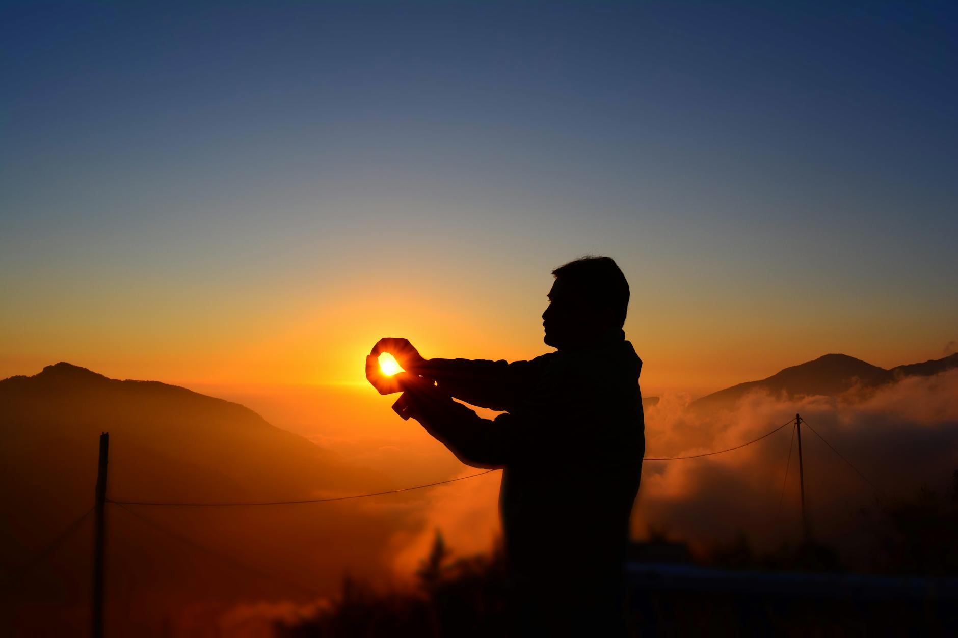 A person silhouetted outdoors against a warm low sunrise over mountains, facing the morning light