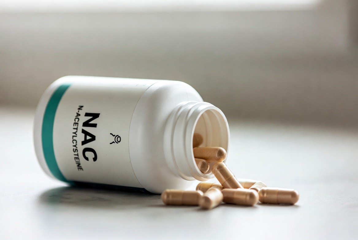 A white supplement bottle with a teal stripe labeled NAC / N-acetylcysteine tipped on its side, with tan oral capsules spilling from the mouth onto a clean light surface &mdash; the over-the-counter dietary-supplement form of the compound