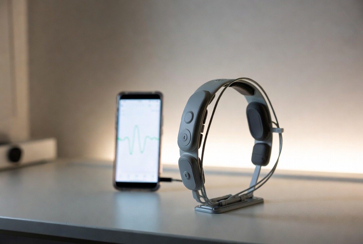 A consumer EEG meditation headband resting on a stand on a calm desk surface beside a soft brainwave visualization on a screen, the headband the clear visual anchor, no identifiable faces