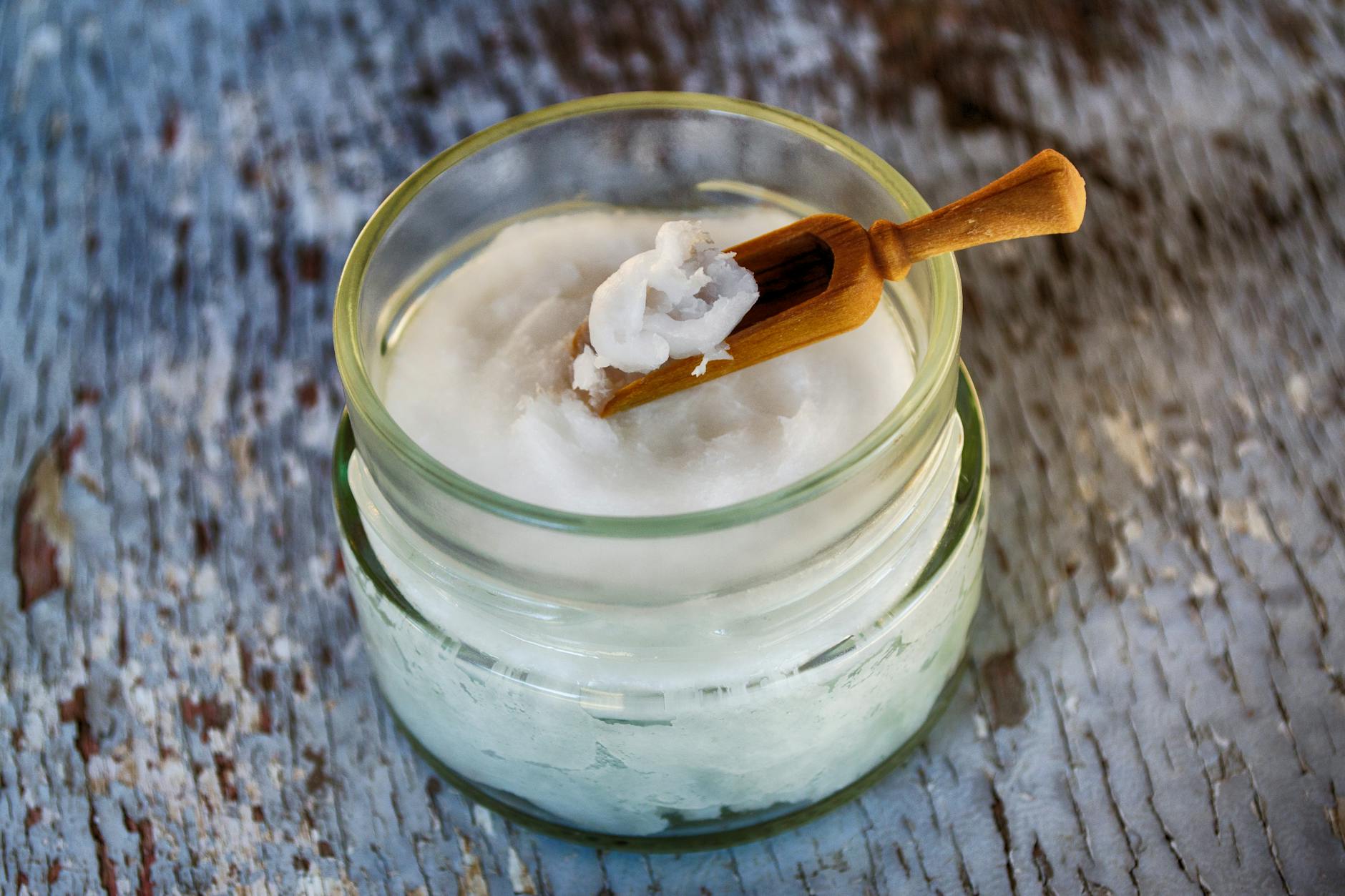 An open glass jar of solid white coconut oil with a wooden scoop resting on the rim, on a weathered counter