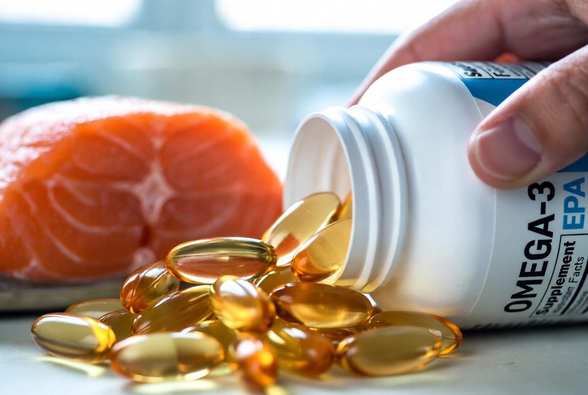 A hand tipping amber omega-3 fish oil softgel capsules from a bottle labeled OMEGA-3 EPA with a raw salmon fillet blurred behind as the natural source in clean kitchen light