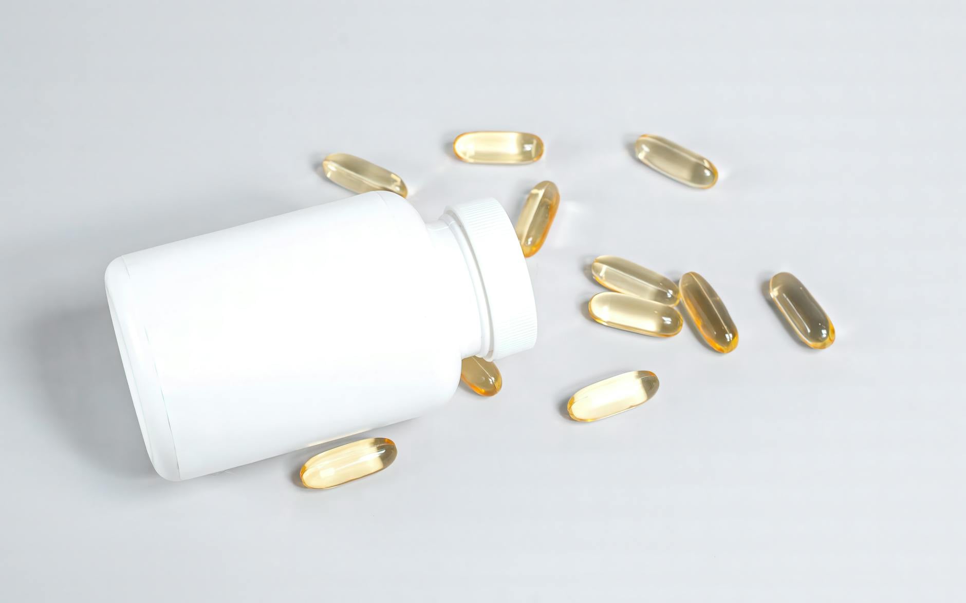 Amber omega-3 fish oil softgel capsules spilling from an unbranded white supplement bottle on a clean light surface