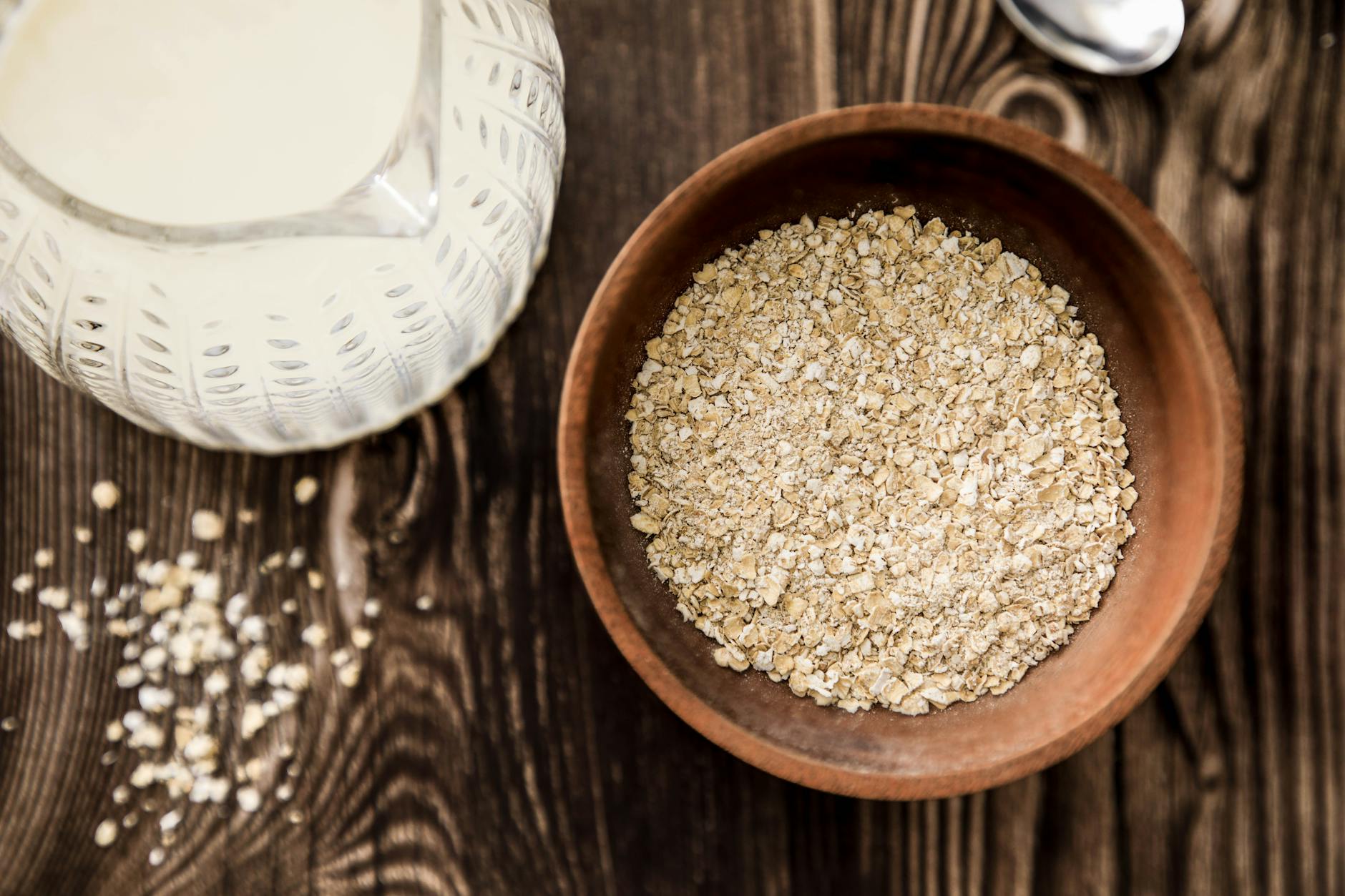 A wooden bowl of rolled oats beside a jug of milk and scattered oat flakes on a dark wood table — oats are a soluble-fiber staple, the same gel-forming fiber family as psyllium