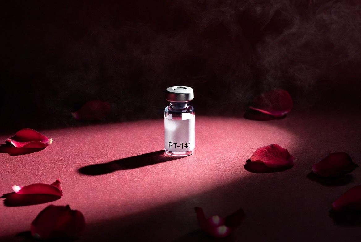 A single glass vial labeled PT-141 (bremelanotide) lit in a soft spotlight on a deep burgundy surface scattered with red rose petals, evoking the libido peptide; no people
