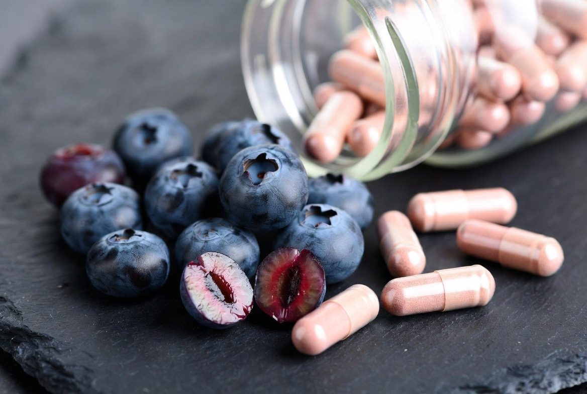 Rose-copper pterostilbene supplement capsules spilling from a glass bottle beside a cluster of fresh ripe blueberries, some sliced open, the natural dietary source of the compound, on a dark slate surface
