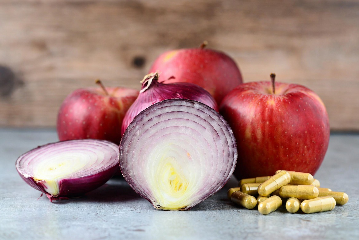 Whole and halved red onions and red apples — among the richest dietary sources of quercetin — beside a small pile of golden supplement capsules on a neutral surface