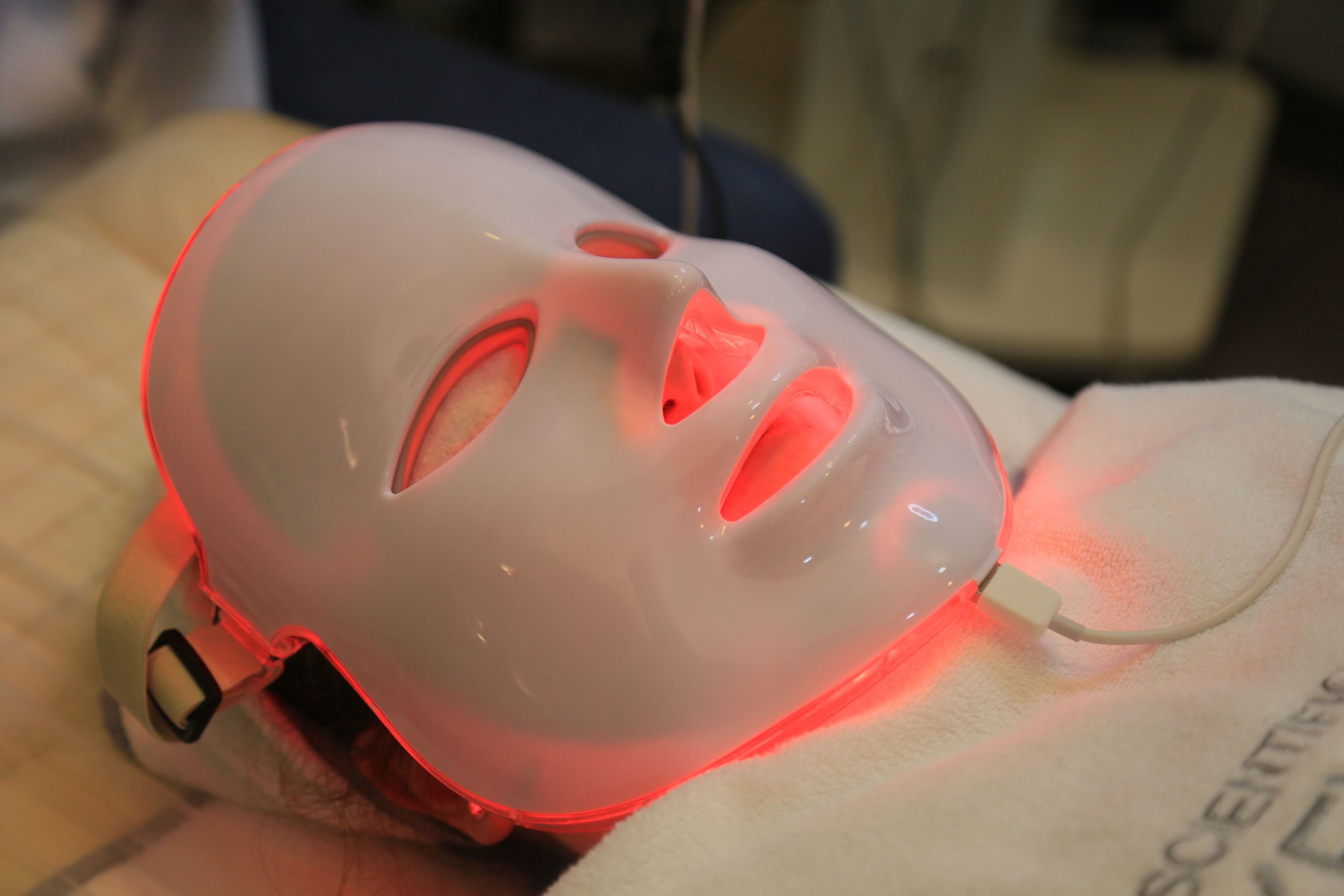 A rigid white at-home LED face mask glowing red while worn, strap and power cable visible, representing a consumer red-light skincare device