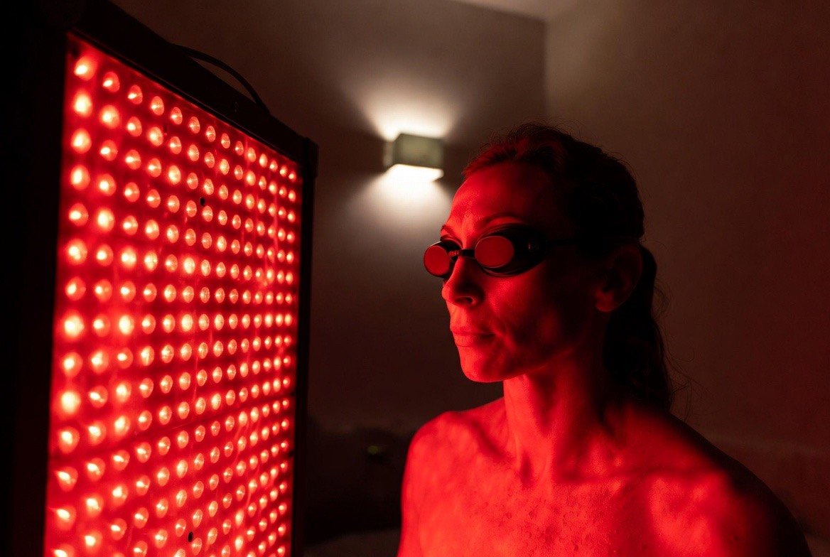 A woman sitting close to a full-body red-light therapy panel in a dim room, her face and torso bathed in deep red LED glow while she wears opaque protective goggles — the everyday at-home photobiomodulation setup where distance and dose decide whether anything happens