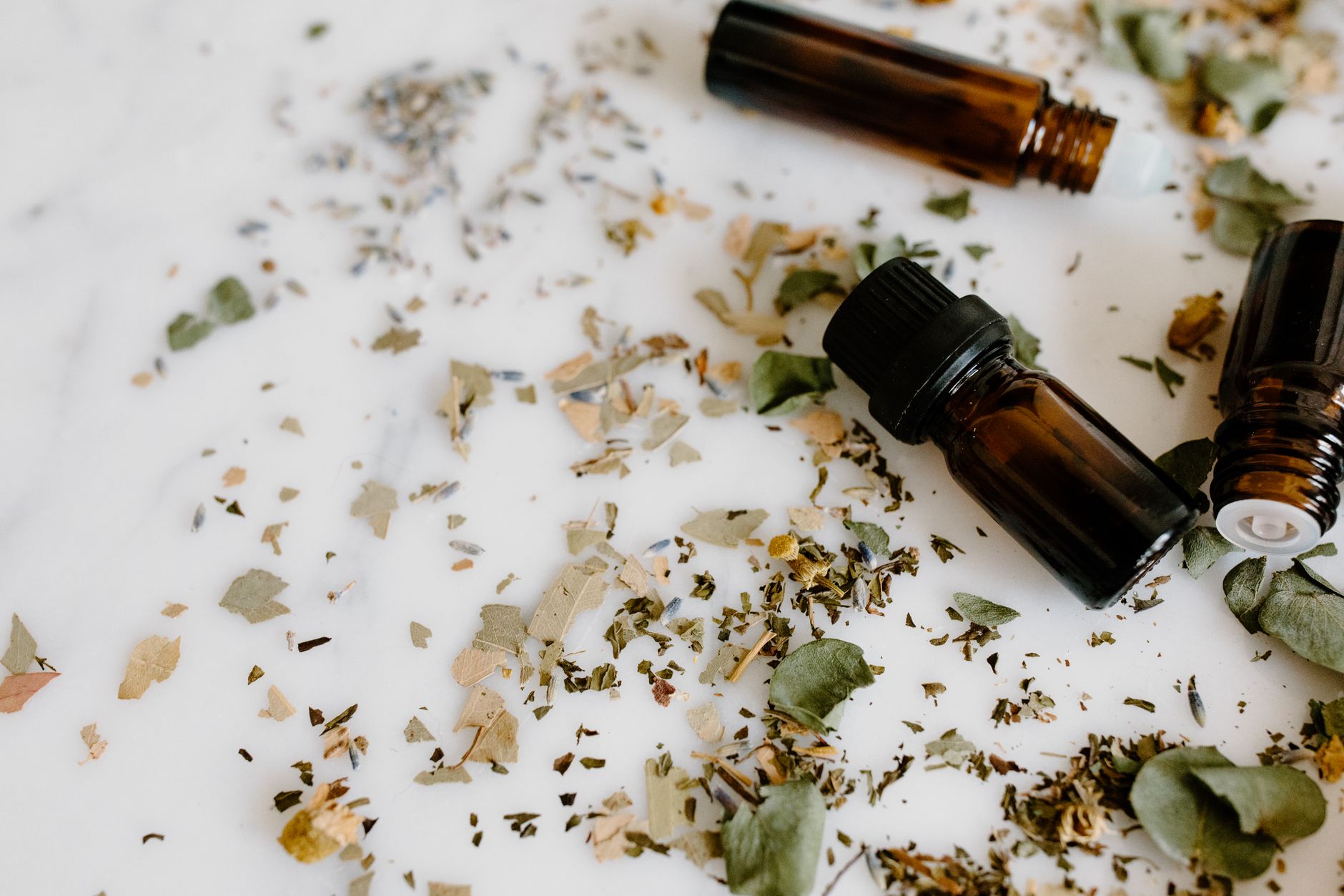 Amber glass dropper bottles of essential oil surrounded by scattered dried herbs and leaves on a white marble surface in soft natural light