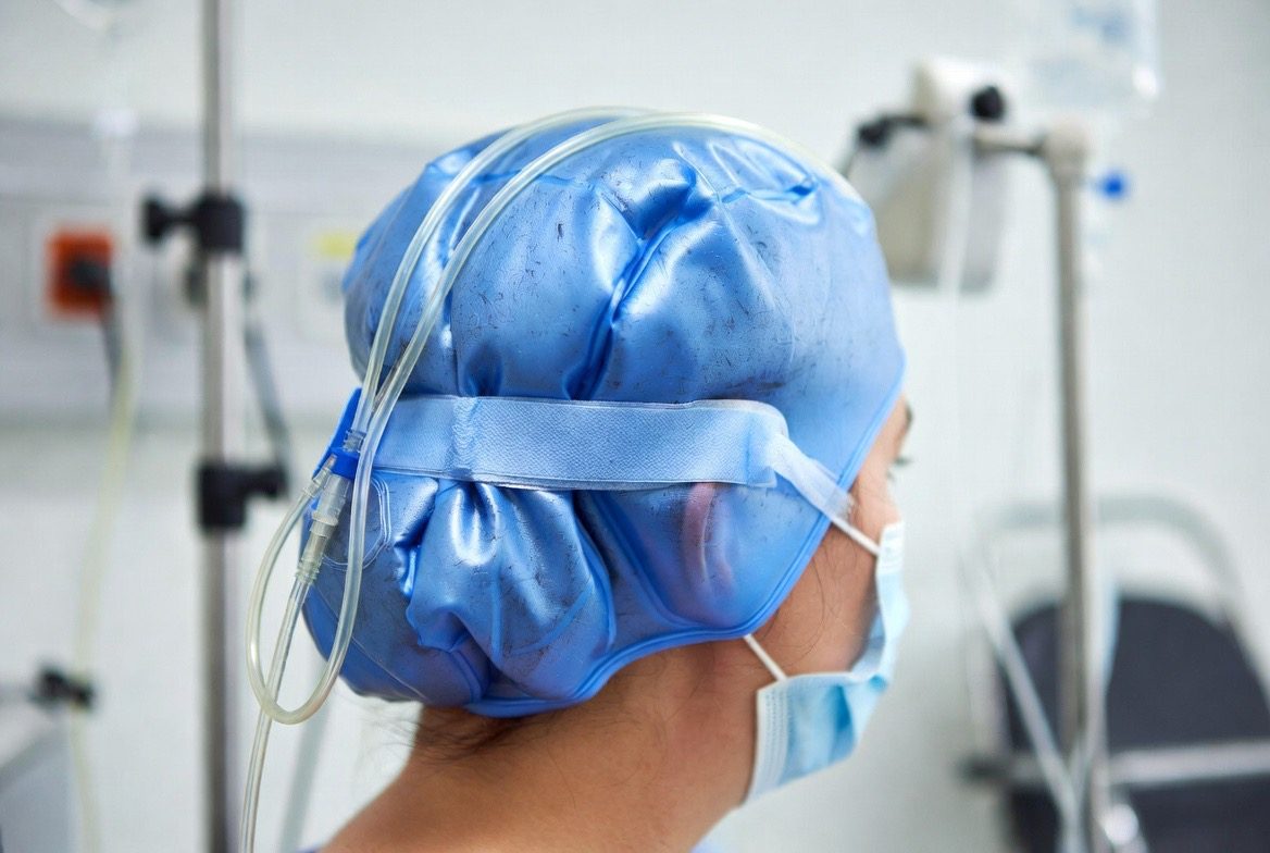 A blue silicone scalp-cooling cold cap worn on a chemotherapy patient's head, connected by insulated cooling tubes, with IV poles behind them during a chemotherapy infusion session to reduce hair loss