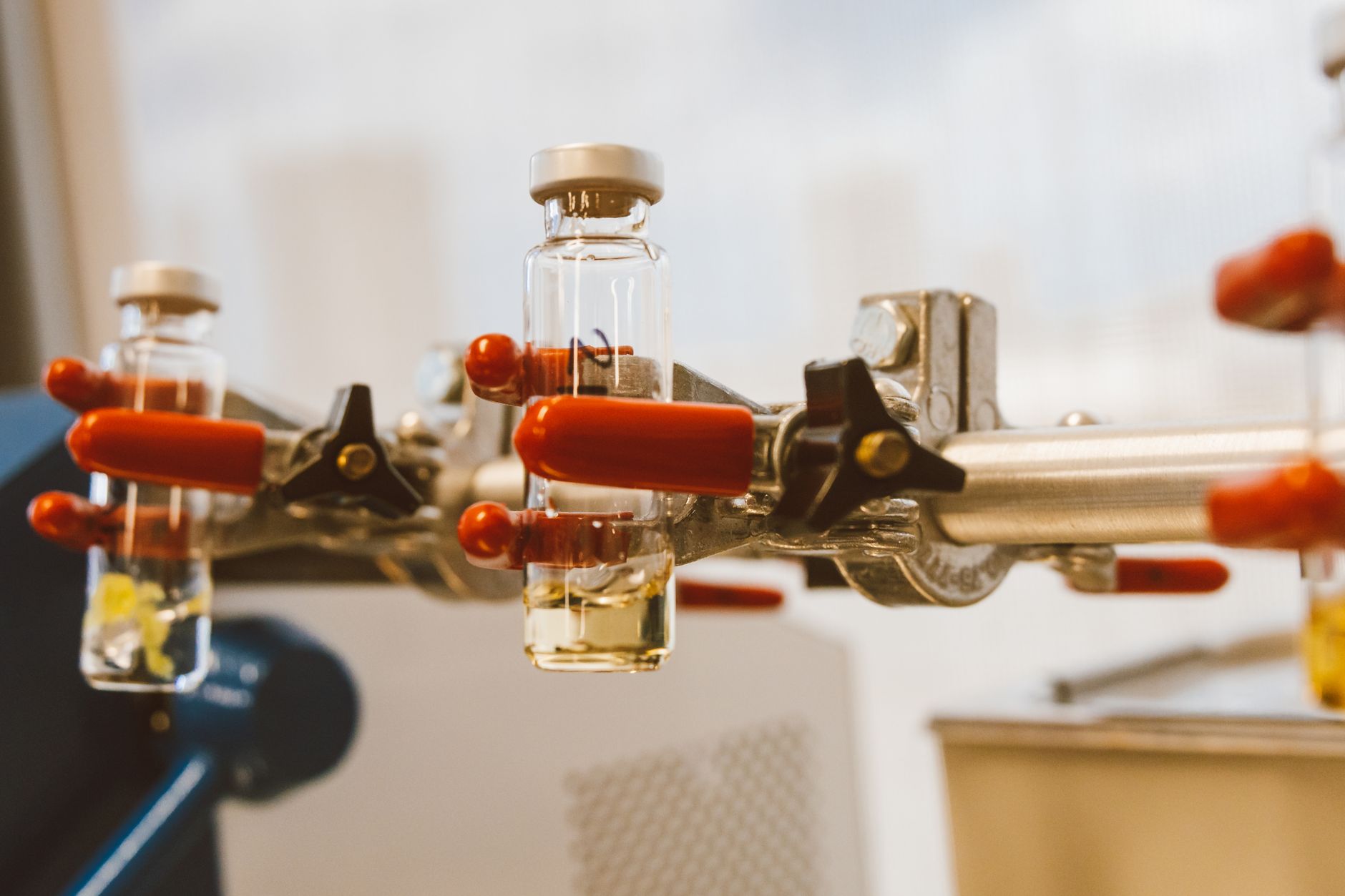 Clear glass research vials held in red-handled laboratory clamps on a stand against a soft clinical background