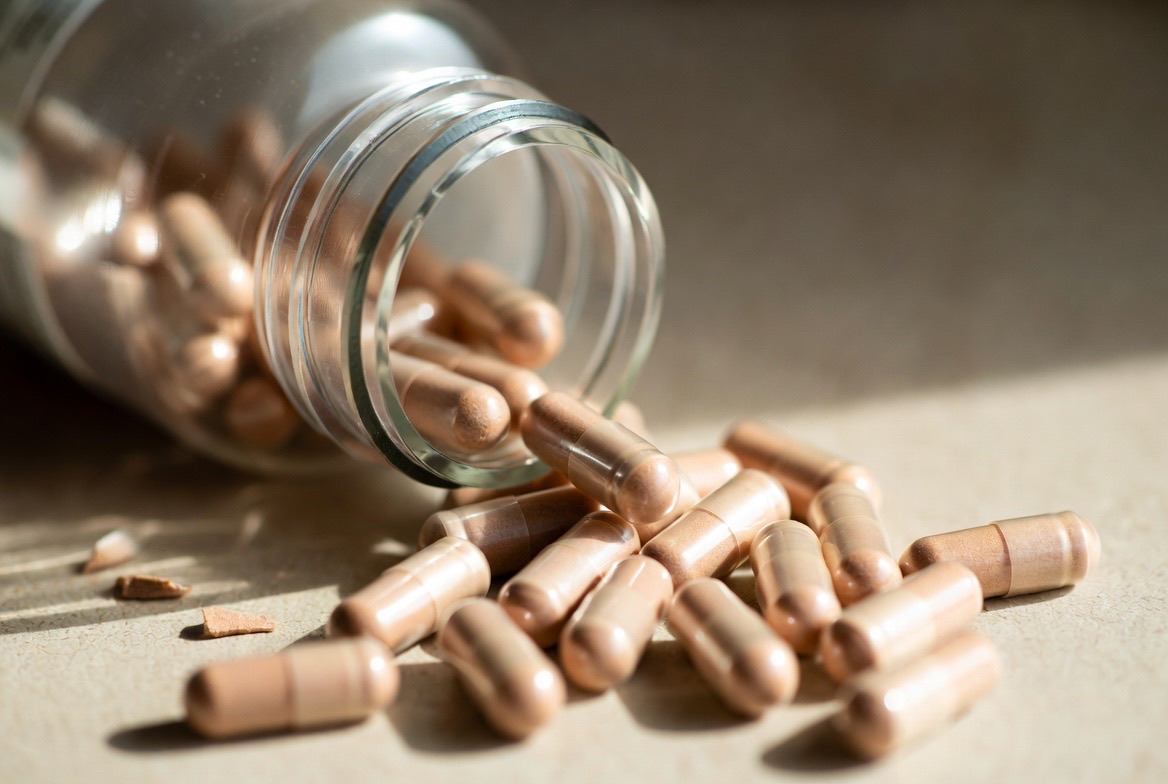 A scattering of clear and white enzyme supplement capsules in and around an open amber bottle on a neutral surface, representing over-the-counter serrapeptase products
