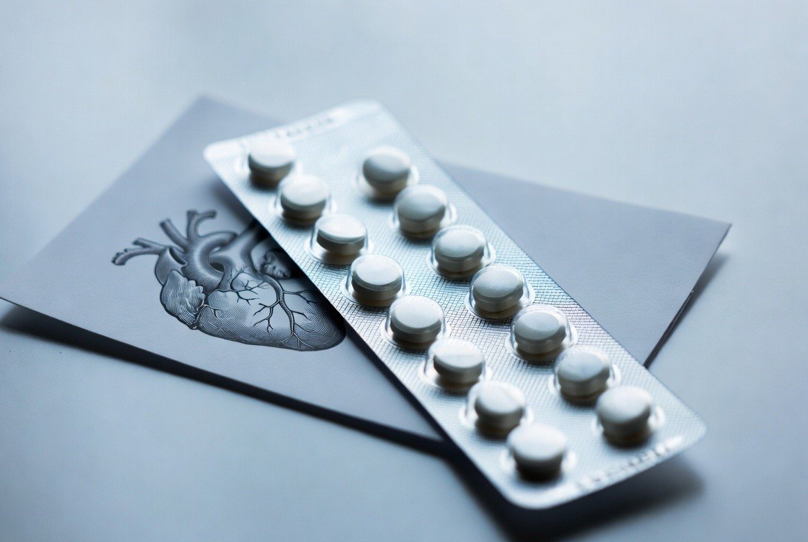 An SGLT2-inhibitor oral tablet blister pack beside a handheld blood-glucose meter and a medical illustration of a heart and kidney, on a clinician's desk