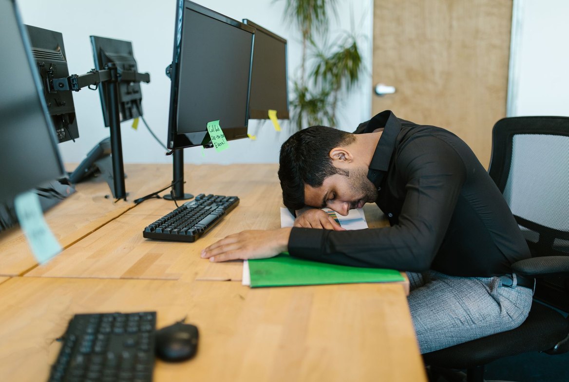 An office worker collapsed head-down on his desk in front of a multi-monitor computer setup, worn out after long hours seated &mdash; the picture of prolonged, uninterrupted sitting that large cohort studies link to higher mortality