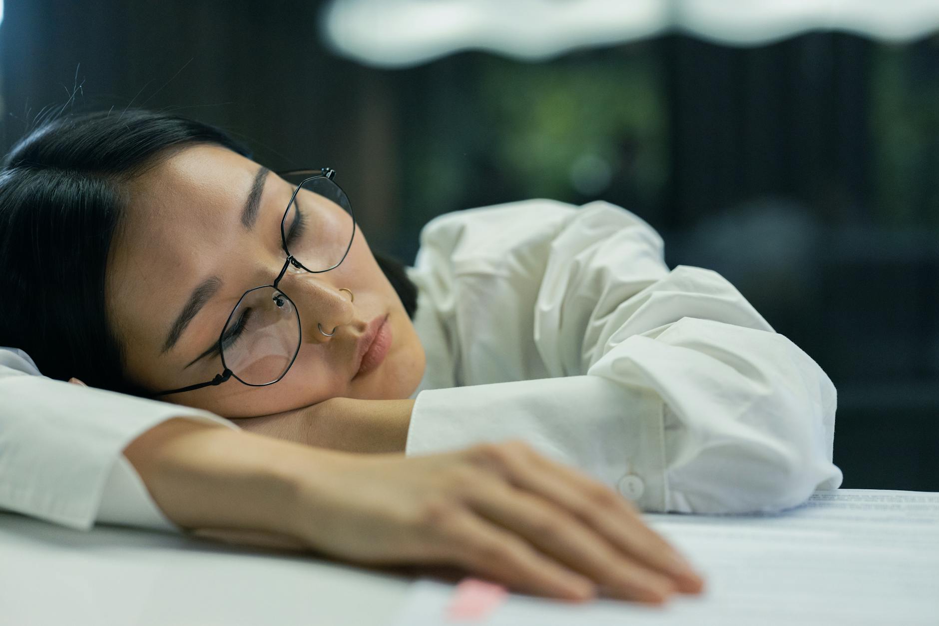 Young woman asleep at a desk with glasses still on — the cumulative cognitive fatigue from chronic sleep debt that Van Dongen 2003, Belenky 2003, and Banks 2010 measured with the psychomotor vigilance task